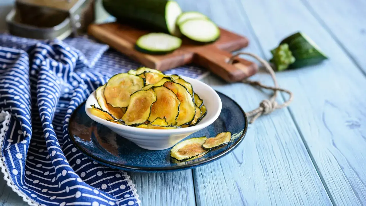 Knusprige Zucchini-Chips in einer Schüssel, daneben frische Zucchinischeiben auf einem Holzbrett.