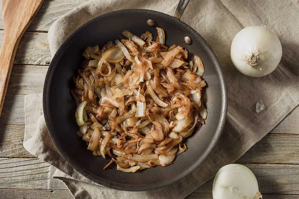 Gebratene Zwiebeln in einer Pfanne, goldbraun und karamellisiert. Nebenbei liegen zwei ganze Zwiebeln und ein Holzspatel. Perfekt zum Zwiebelgrillen!
