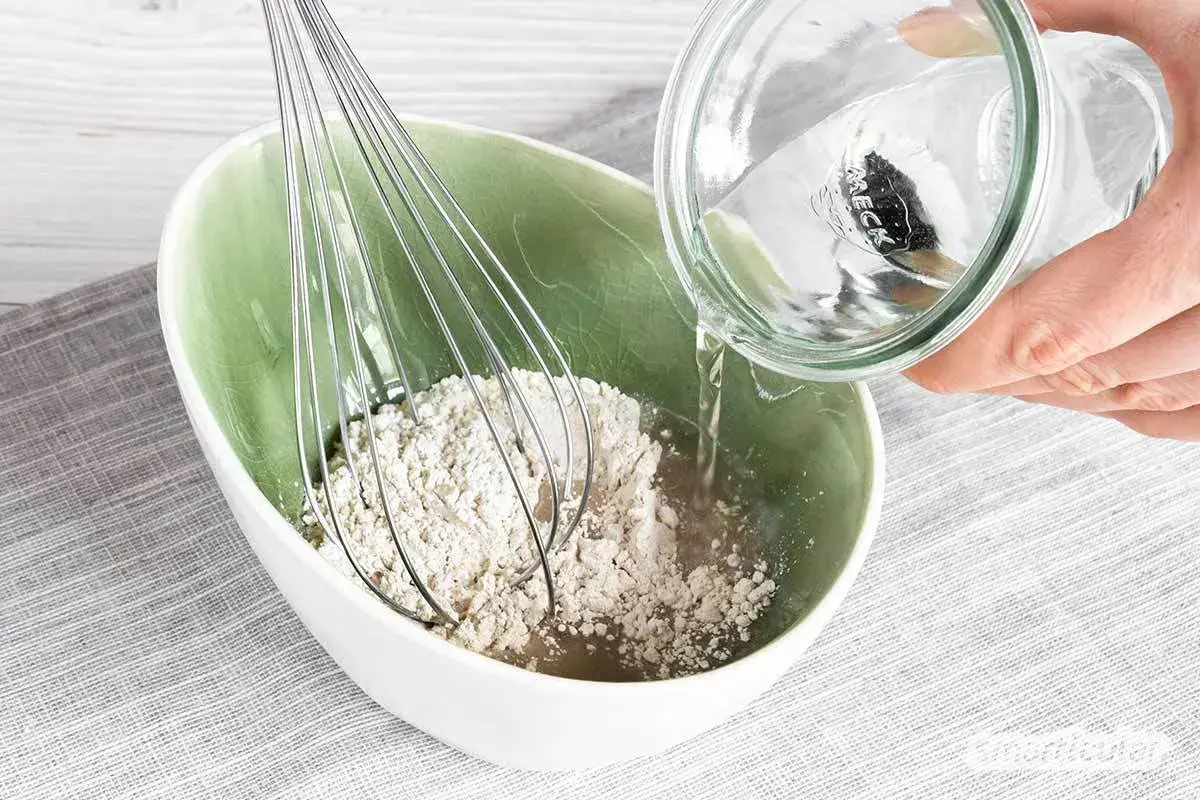 Eine Hand gießt Wasser aus einem Glas in eine Schüssel mit Roggenmehl und einem Schneebesen. Ein natürliches Shampoo aus Roggenmehl wird zubereitet.