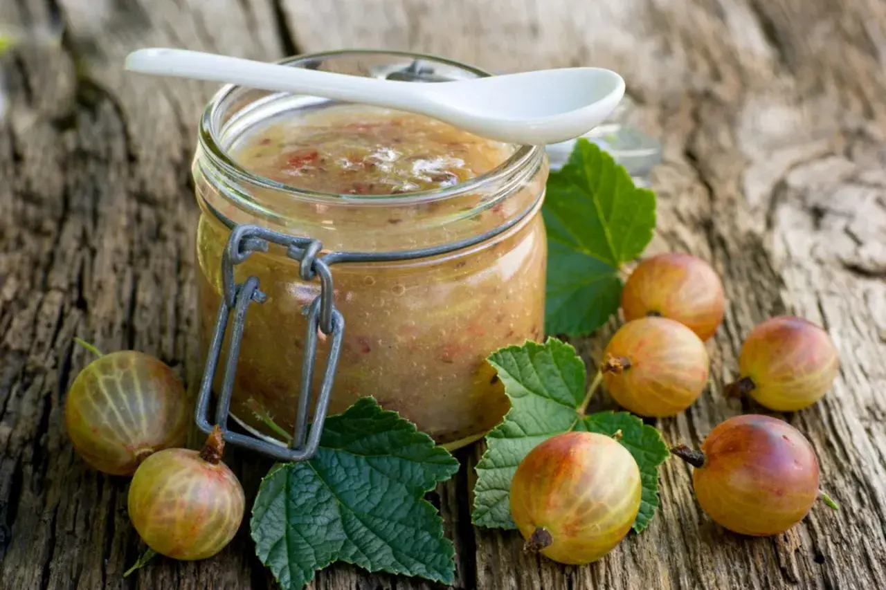 Glas mit selbstgemachter Stachelbeer Marmelade, umgeben von frischen Stachelbeeren und Blättern auf rustikalem Holz.