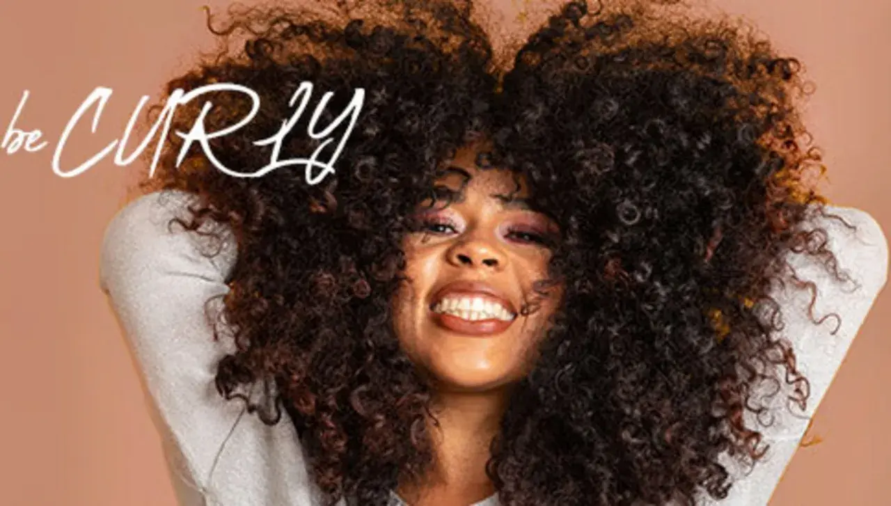 Frau mit vollem, lockigem Haar, die lächelt. Der Schriftzug "be CURLY" ist im Bild zu sehen.