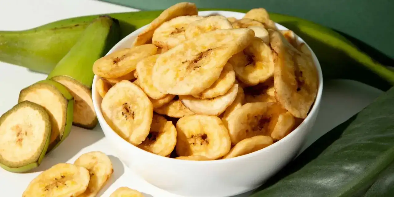 Schmackhafte bananenchips selber machen ist ganz einfach. Hier sind sie in einer Schüssel, daneben grüne Bananen und Blätter.