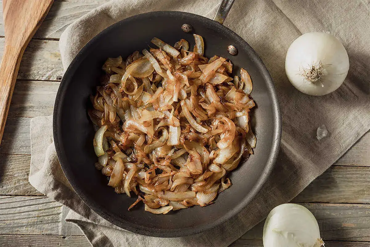 Gebratene Zwiebeln in einer Pfanne, goldbraun und karamellisiert. Nebenbei liegen zwei ganze Zwiebeln. Perfekt zum Zwiebelgrillen!