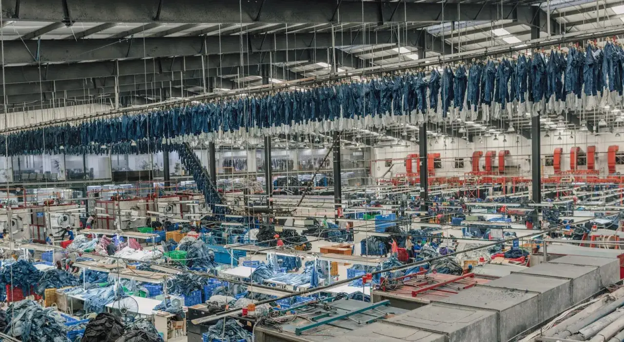 Gro&szlig;e Fabrikhalle mit Reihen von aufgeh&auml;ngten Jeans, die auf den Abschluss der Jeans Herstellung warten. Unten sind Arbeitsstationen mit Stoffen und Maschinen.