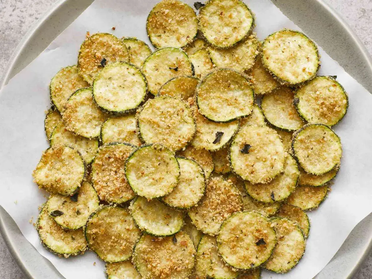 Knusprige Zucchini-Chips, goldbraun gebacken und mit Kräutern bestreut, liegen auf Backpapier.
