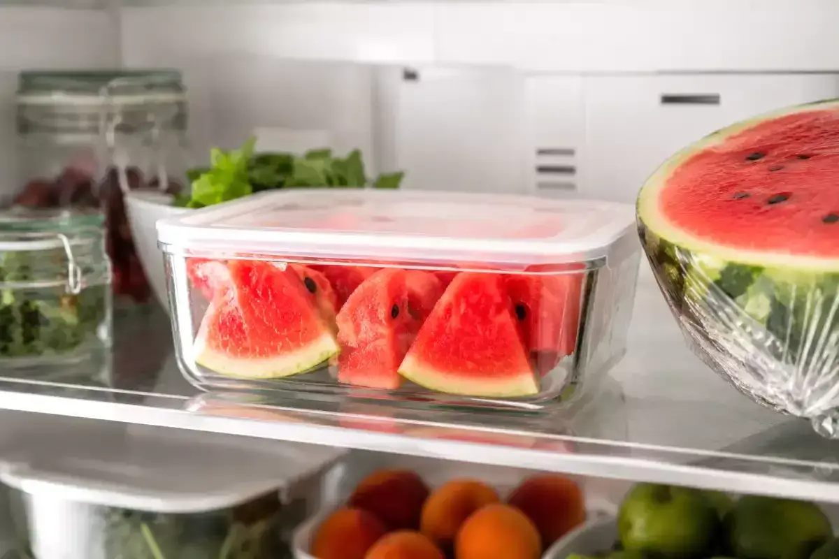 Wassermelone in Scheiben im Kühlschrank, um die Haltbarkeit zu verlängern. Daneben eine halbe Wassermelone in Folie.