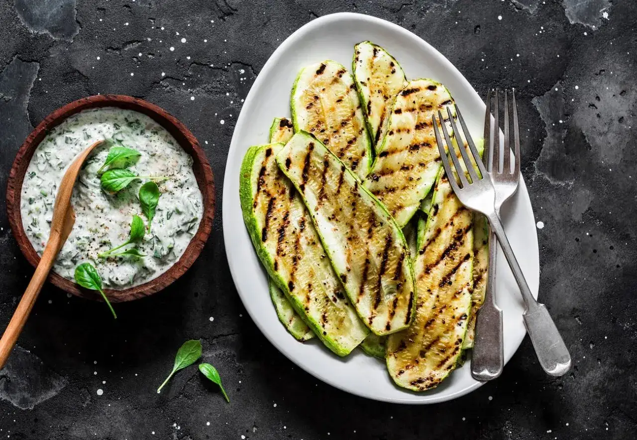Perfekt gegrillte Zucchini-Scheiben auf einem Teller, daneben eine Schüssel mit cremigem Dip und frischen Kräutern. Ein Genuss für jeden Grillabend!