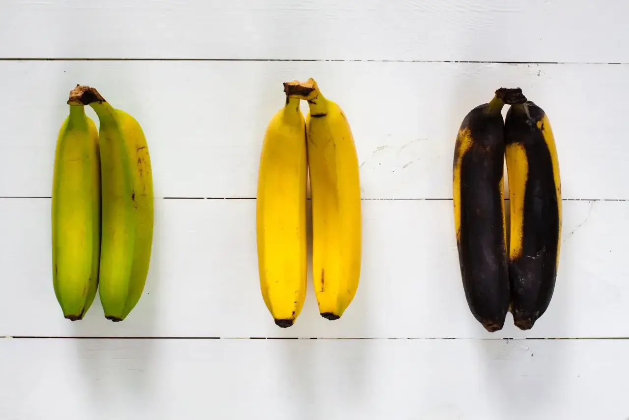Drei Paare Bananen auf wei&szlig;em Holz: gr&uuml;n, gelb und &uuml;berreif. So kann man Bananen lagern, um den Reifeprozess zu beobachten.
