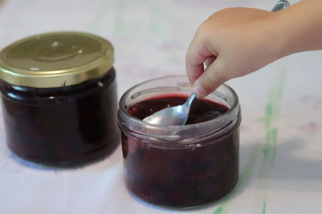 Ein Kinderfinger stochert mit einem Löffel in einem Glas mit Zwetschgen zum Einkochen.