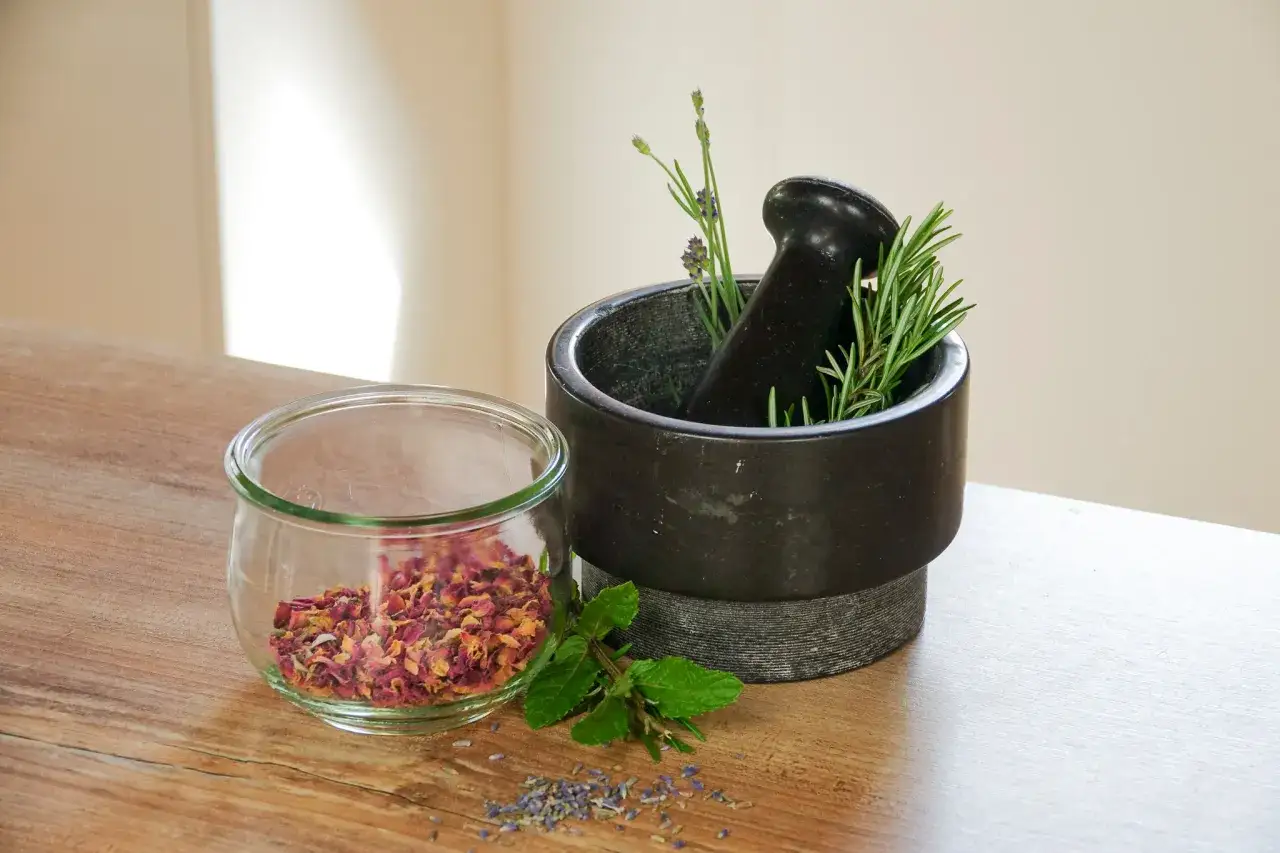 Glas mit Rosenblättern, Mörser mit Kräutern wie Rosmarin und Lavendel – Zutaten für kosmetik selber machen.