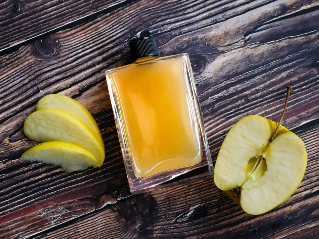 Apfelessig in einer Flasche, daneben Apfelschnitze und eine Apfelh&auml;lfte. Ideal f&uuml;r die Hautpflege.