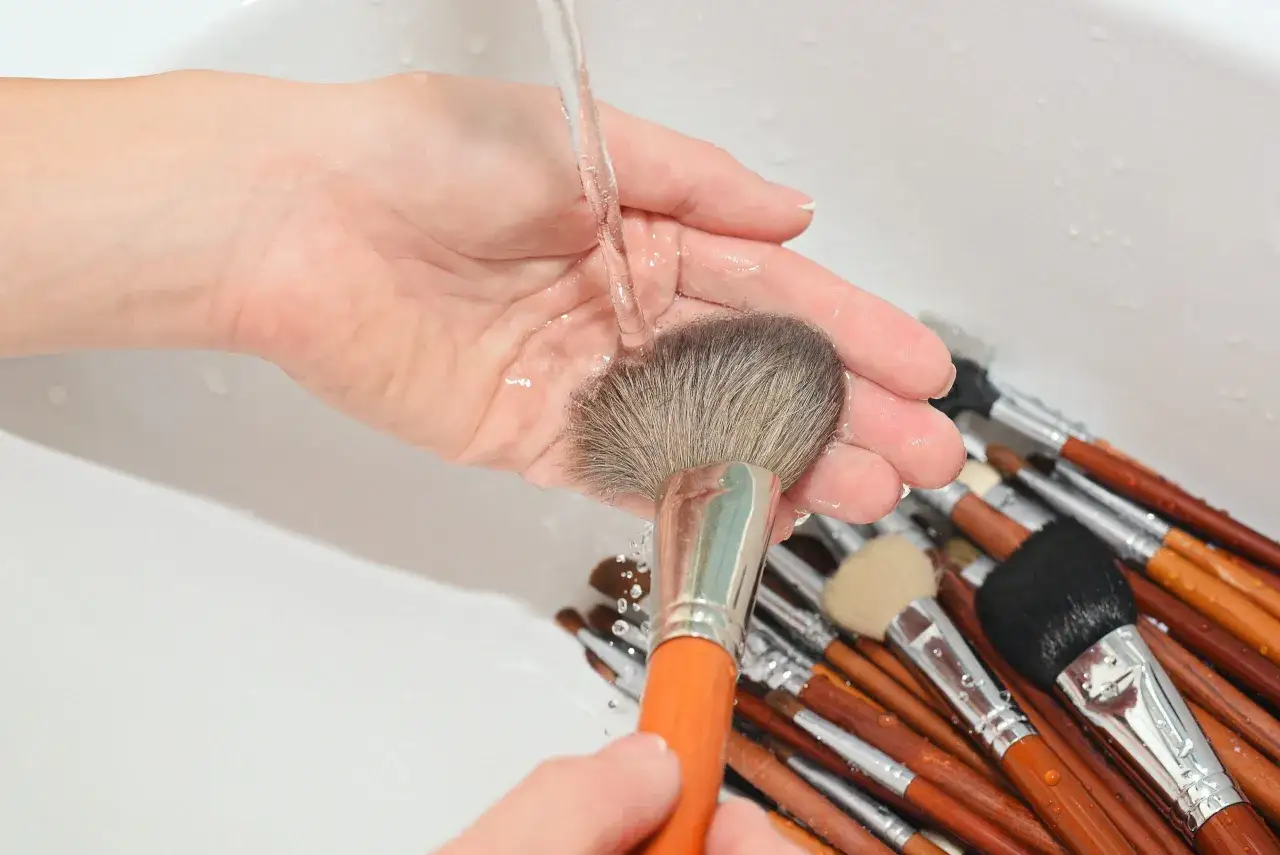 Eine Hand hält einen Schminkpinsel unter fließendem Wasser, um ihn zu reinigen. Viele weitere Pinsel liegen in der Spüle.