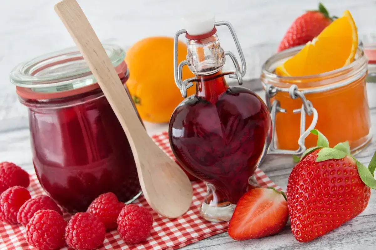 Frische Beeren und Orangen laden zum Marmelade kochen ein. Ein Glas mit roter Marmelade, eine Herzflasche mit Sirup und ein Glas mit Orangenmarmelade stehen bereit.