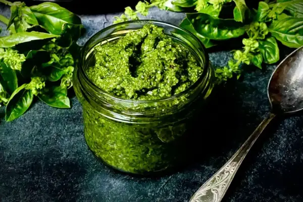 Ein Glas mit selbstgemachtem Pesto, umgeben von frischem Basilikum. Daneben liegt ein Löffel.