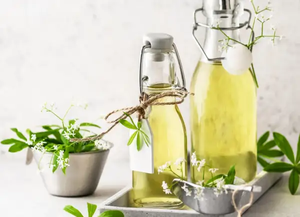 Zwei Flaschen mit hellem, gelbem Waldmeister Sirup, verziert mit frischen Waldmeisterzweigen und kleinen weißen Blüten.