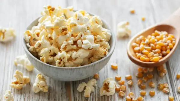 Schüssel mit frisch gemachtem Popcorn, daneben eine Holzschaufel mit Maiskörnern. So einfach ist Popcorn selber machen!
