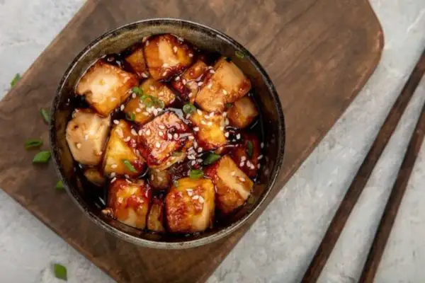 Ofen-gebackener Tofu in einer dunklen, glänzenden Sauce, bestreut mit Sesam und Frühlingszwiebeln, serviert in einer Schüssel auf einem Holzbrett.