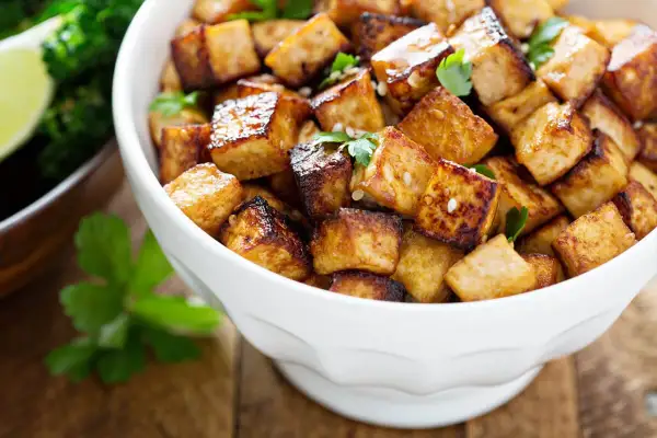 Knusprig gebratene Tofuwürfel, perfekt zum tofu zubereiten, garniert mit Petersilie und Sesam. Daneben ein Schälchen mit Brokkoli und Limette.