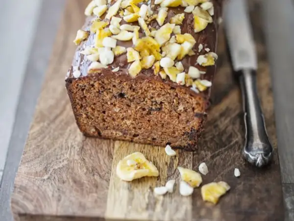 Saftiger bananenkuchen vegan mit Schokoladenglasur und Nüssen, auf einem Holzbrett mit einem Messer.