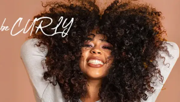 Frau mit vollem, lockigem Haar, die lächelt. Der Schriftzug "be CURLY" ist im Bild zu sehen.