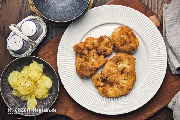 Knusprig **gebratener Blumenkohl** auf einem weißen Teller, daneben Kartoffelsalat und ein Salz- und Pfefferstreuer.