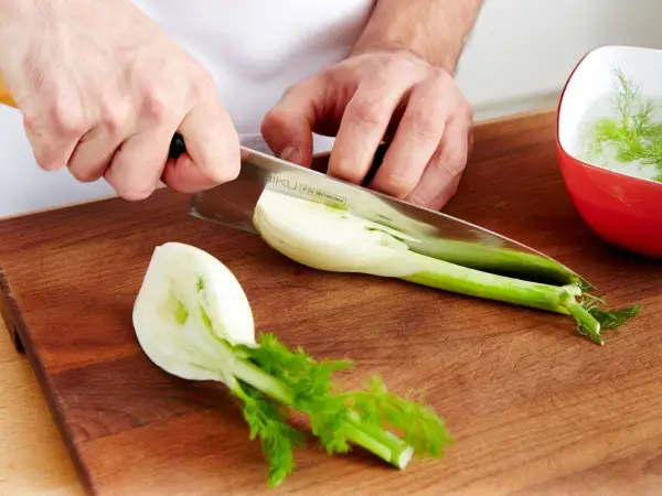 Hände schneiden frischen Fenchel für den Verzehr. Ein Teil liegt auf dem Holzbrett, ein anderer in einer roten Schüssel mit Wasser.